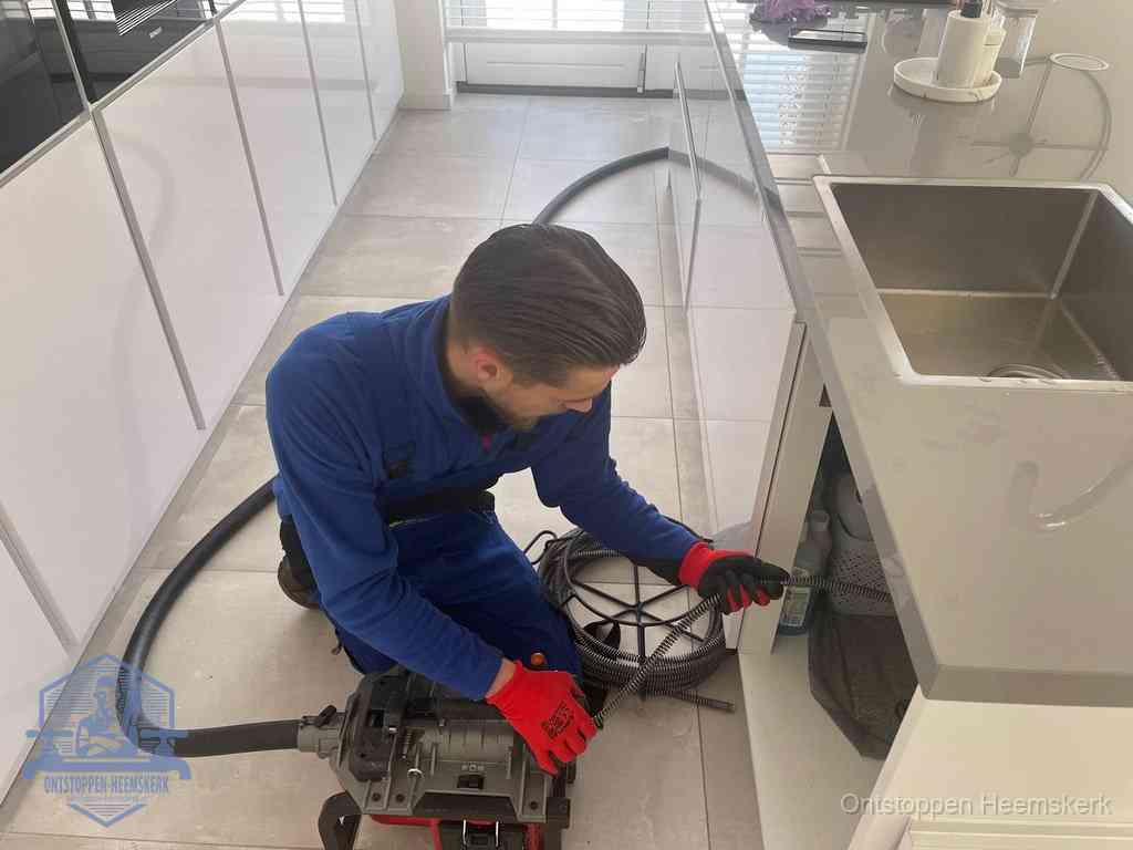 Een monteur ontstopt de keukenafvoer onder de spoelbak met een elektrische veermachine in Heemskerk