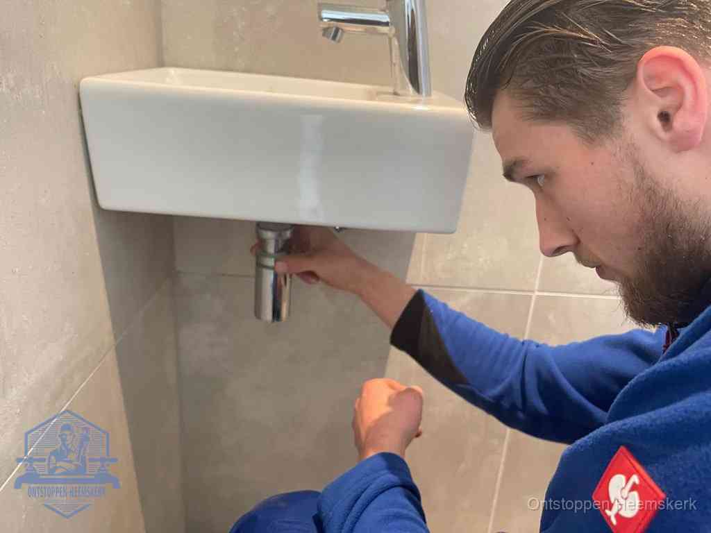 Een monteur draait met de hand de zwanenhalssifon onder een fonteinwasbak in de toiletruimte los om de bron van rioollucht in Heemskerk te vinden